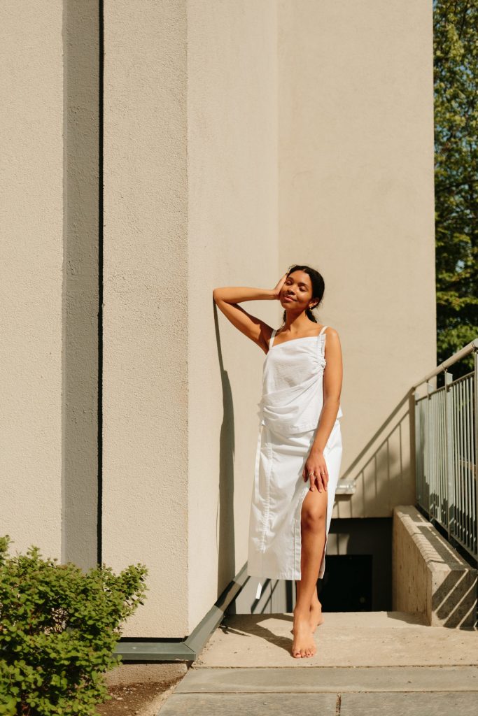 totalbilde av en kvinnelig modell iført en løs, hvit topp og skjørt. Hun lener seg, på en arm, mot en vegg.