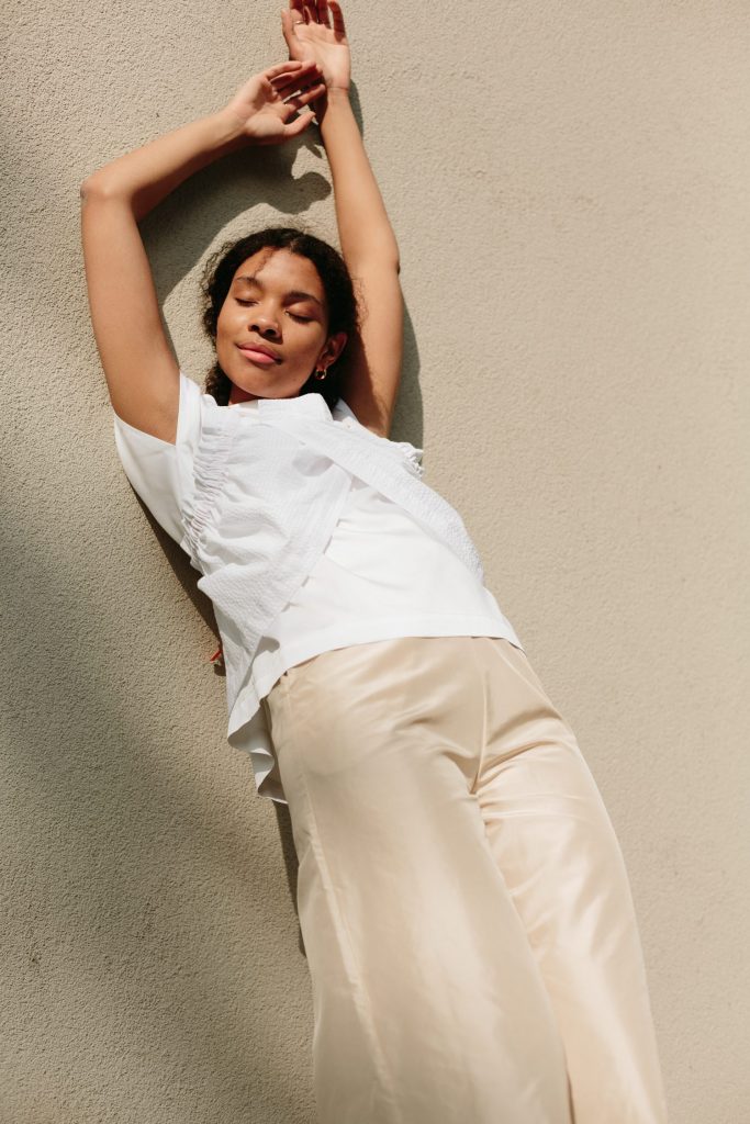 kvinnelig modell iført en beige bukse og en hvit topp lener seg mot en vegg