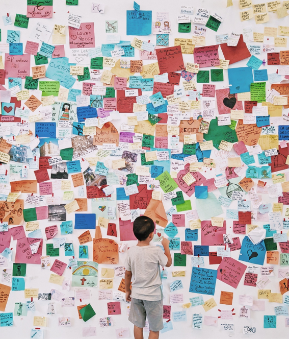 child standing in front of a wall with many drawings and notes.