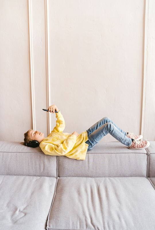 picture of a child laying on a couch with headphones looking at their phone.