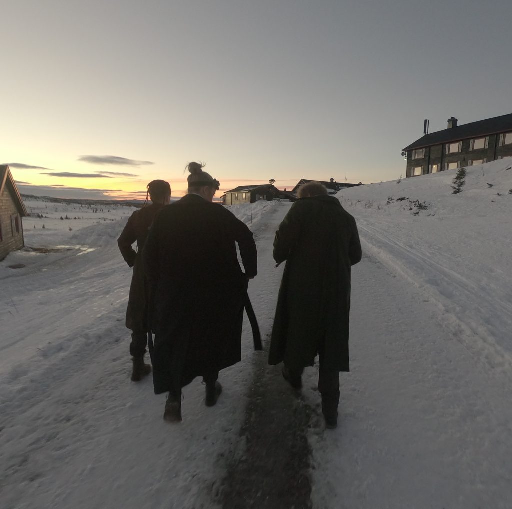 Morgengry, tre personer på en vei i et snødekt landskap, noen hus langs veien.