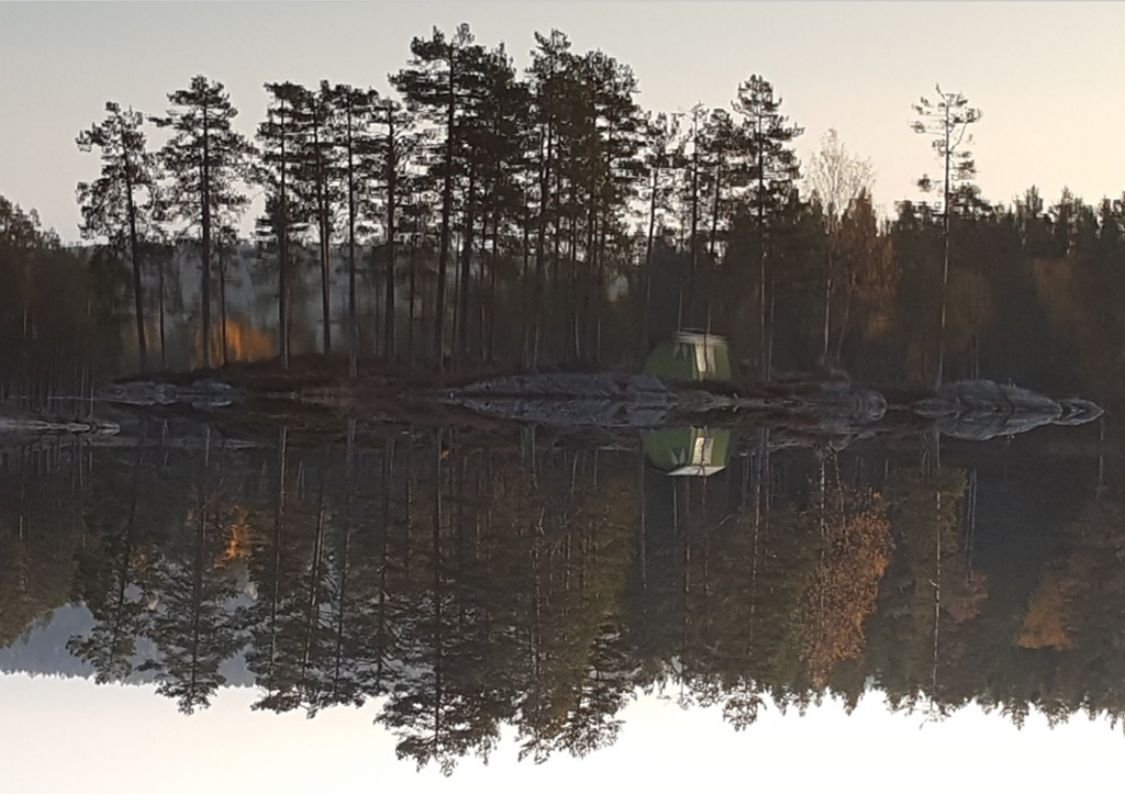 Skoglandskap med teltplass på en øy, trærne speiler seg i innsjøen. 