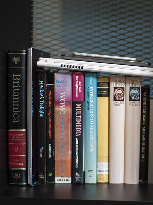 Nærbilde av bokhylle med en blanding av mediefaglige bøker, nettbrett, mobil og PC.