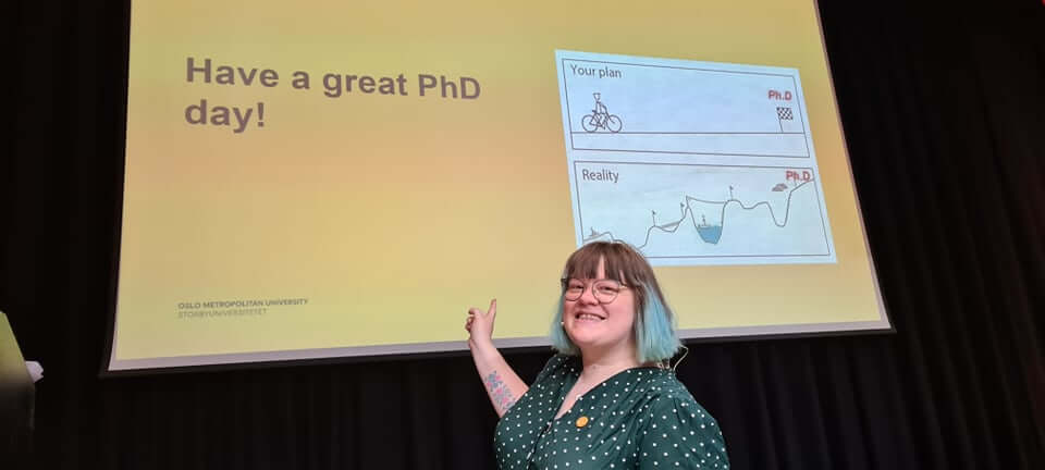 PhD forum leader Camilla Holm smiles and points at screen that reads: Have a great PhD day!