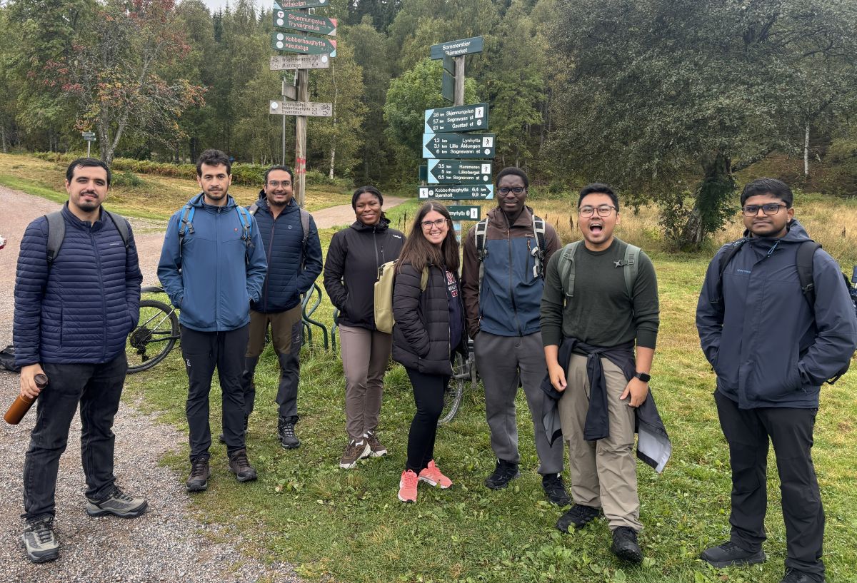 Group of eight RISE members in outdoor hiking gear standing together and smiling on a gravel path. There are wooden trail signposts behind them, showing directions and distances. The group is surrounded by forest and green grass.