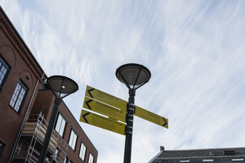 Skiltstolpe med gule retningsskilt som viser vei til ulike bygninger ved OsloMet, omgitt av murbygninger og blå himmel.