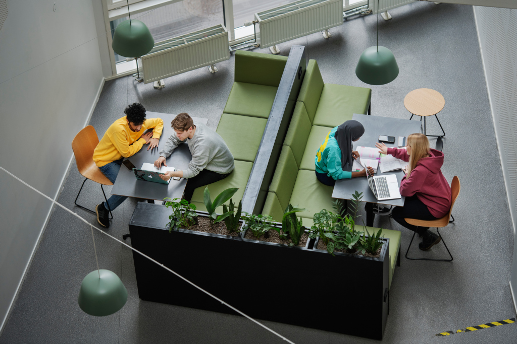 Fire studenter sitter i grupper ved to bord i en moderne campus-lounge og jobber sammen med bøker og bærbare datamaskiner.