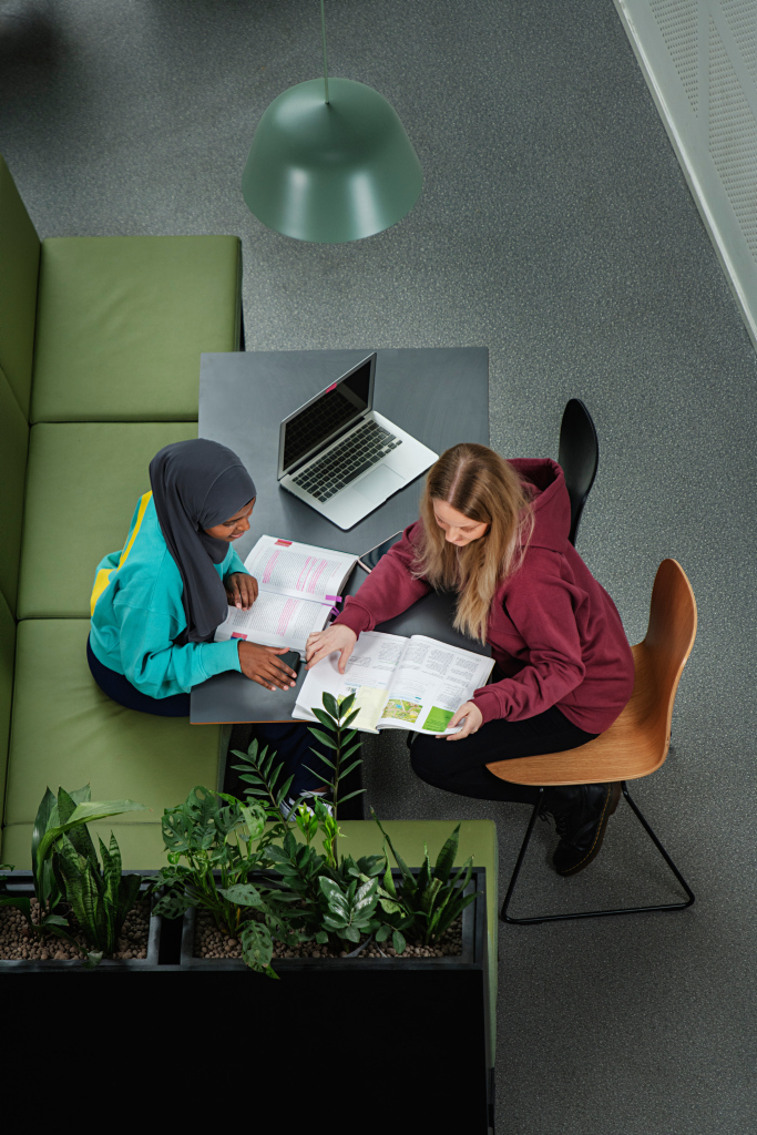 To studenter sitter ved et bord og samarbeider om oppgaver med bøker og bærbar PC i et moderne campusområde.