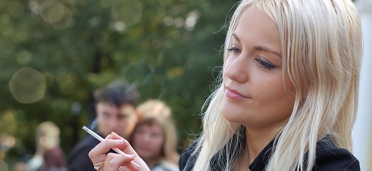Ung blond kvinner røyker ute med folk i bakgrunn. Illustrasjonsfoto: colourbox