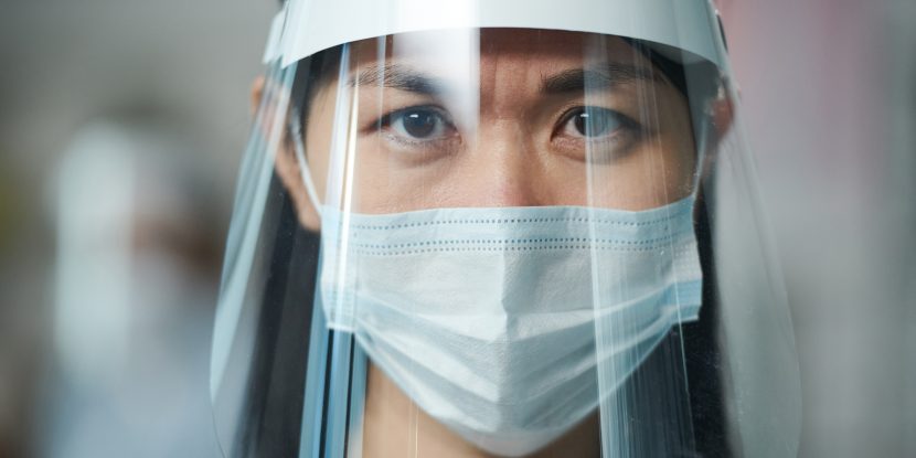 Young serious nurse in uniform and protective workwear looking at you through transparent screen